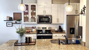 Una cocina moderna con armarios blancos, un refrigerador de acero inoxidable, un microondas y una estufa a gas. Dos lámparas colgantes iluminan la encimera.