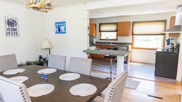 A dining area with a dark wooden table, beige chairs, and placemats. A chandelier hangs from the ceiling, and there are kitchen cabinets and appliances in the background.