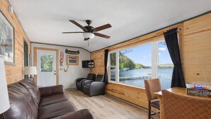 Une cabane avec un canapé en cuir marron, un ventilateur de plafond, une télévision et une fenêtre donnant sur un lac.