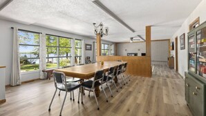 Uma sala de jantar espaçosa com uma longa mesa de madeira e cadeiras, amplas janelas com cortinas, e uma área de cozinha com parede de tijolos e armários de madeira.