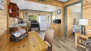 A compact kitchen with a stove, oven, and microwave, wooden walls, a dining table with chairs, and a small seating area with a lamp. - Cozy Cottage Steps From the Water (Emily)