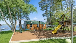 A playground with swings, a slide, and a small wooden house. - Cozy Cottage Steps From the Water (Emily)