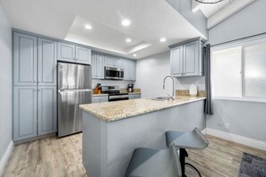 A modern kitchen with grey cabinets, stainless steel appliances, a granite countertop, and a bar area with two stools. - Oceanfront Triplex with Breathtaking Views (Newport Beach)