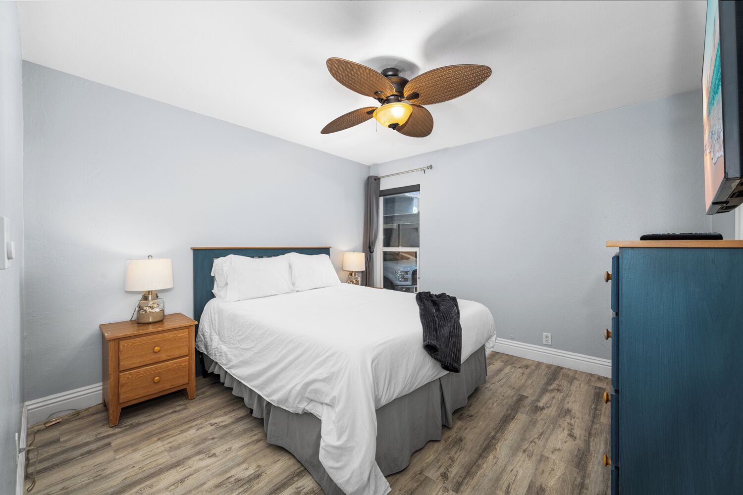 Une chambre à coucher comprenant un lit, des tables de chevet, un ventilateur de plafond, une commode et une fenêtre avec des stores.