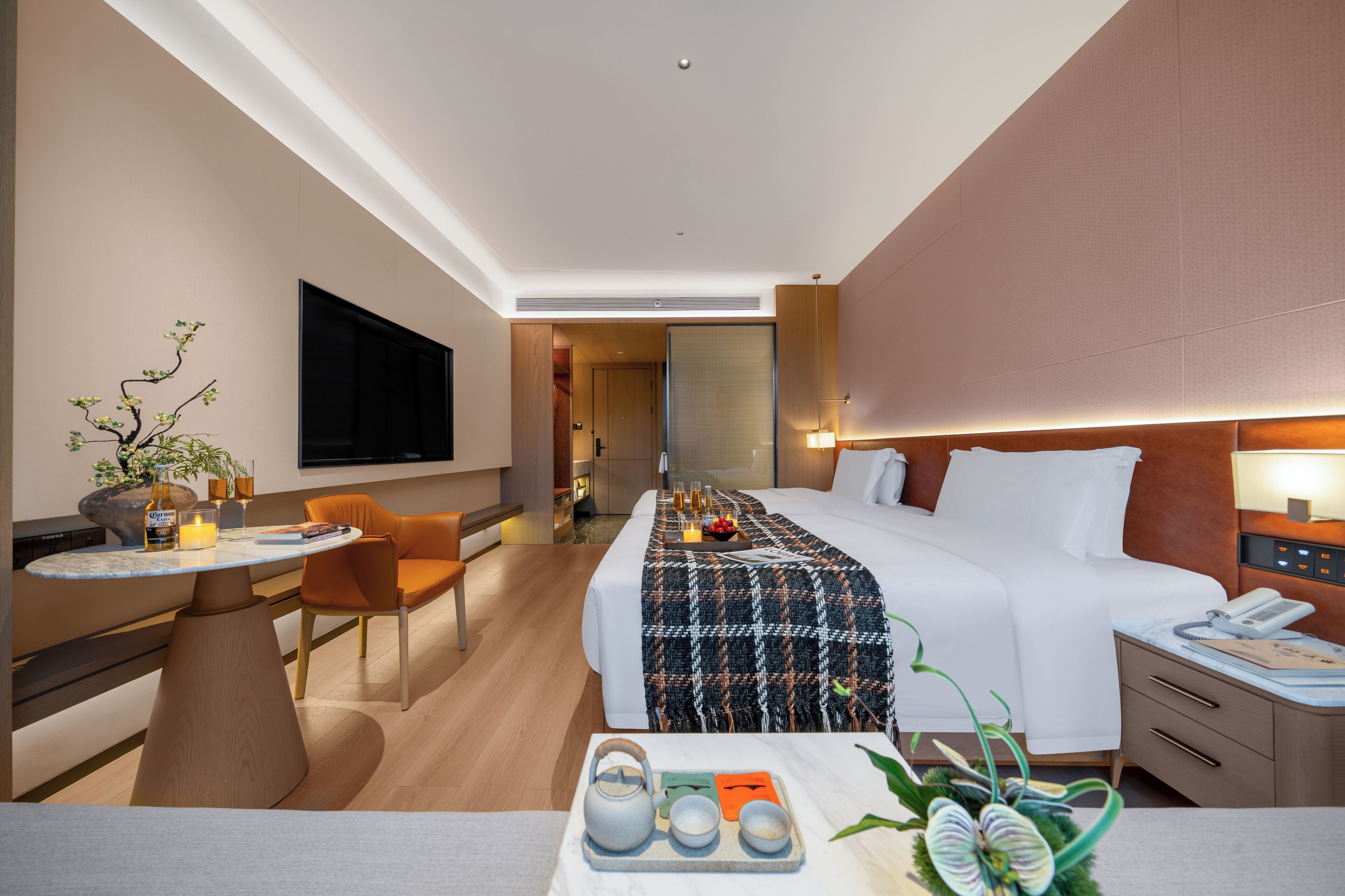 A modern hotel room with a bed, bedside table, chair, and a small table with a tray of tea and snacks.