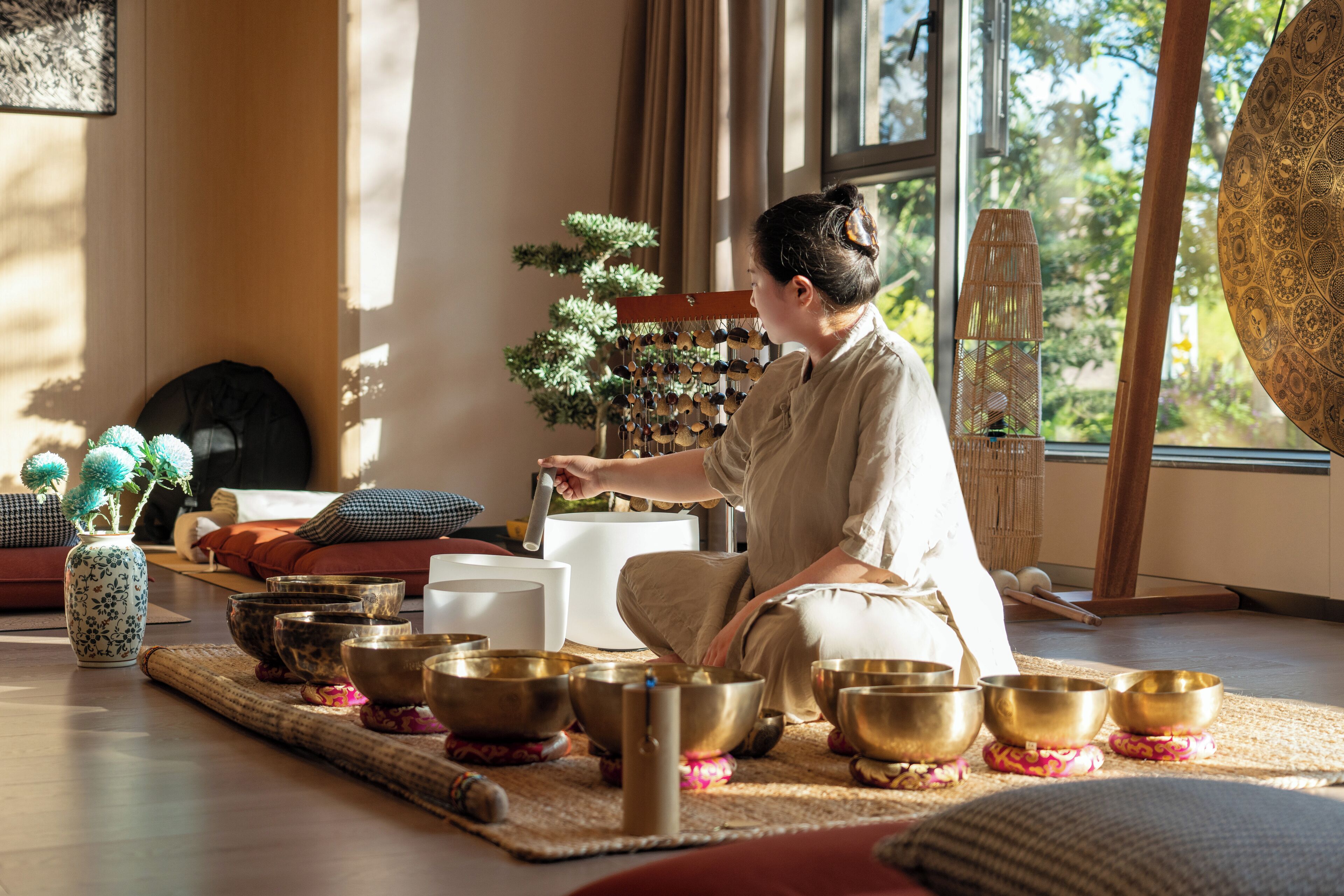 A person sitting on a mat surrounded by multiple bowls, likely used for meditation or sound therapy, in a room with large windows and a view of greenery outside.