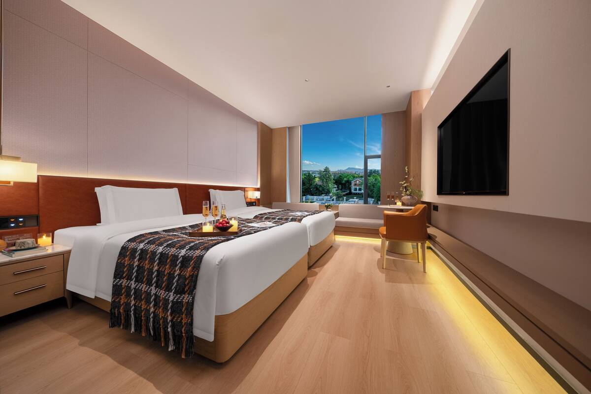 A modern hotel room with two beds, a TV, a bedside table with a lamp, and a view of the city.