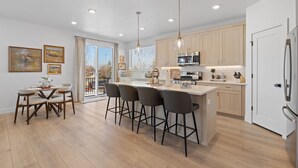 A modern kitchen with wooden cabinets, a central island with bar stools, and a dining area with a table and chairs. - Ski Basecamp • Cozy Modern • Minutes to Everything (Midvale)
