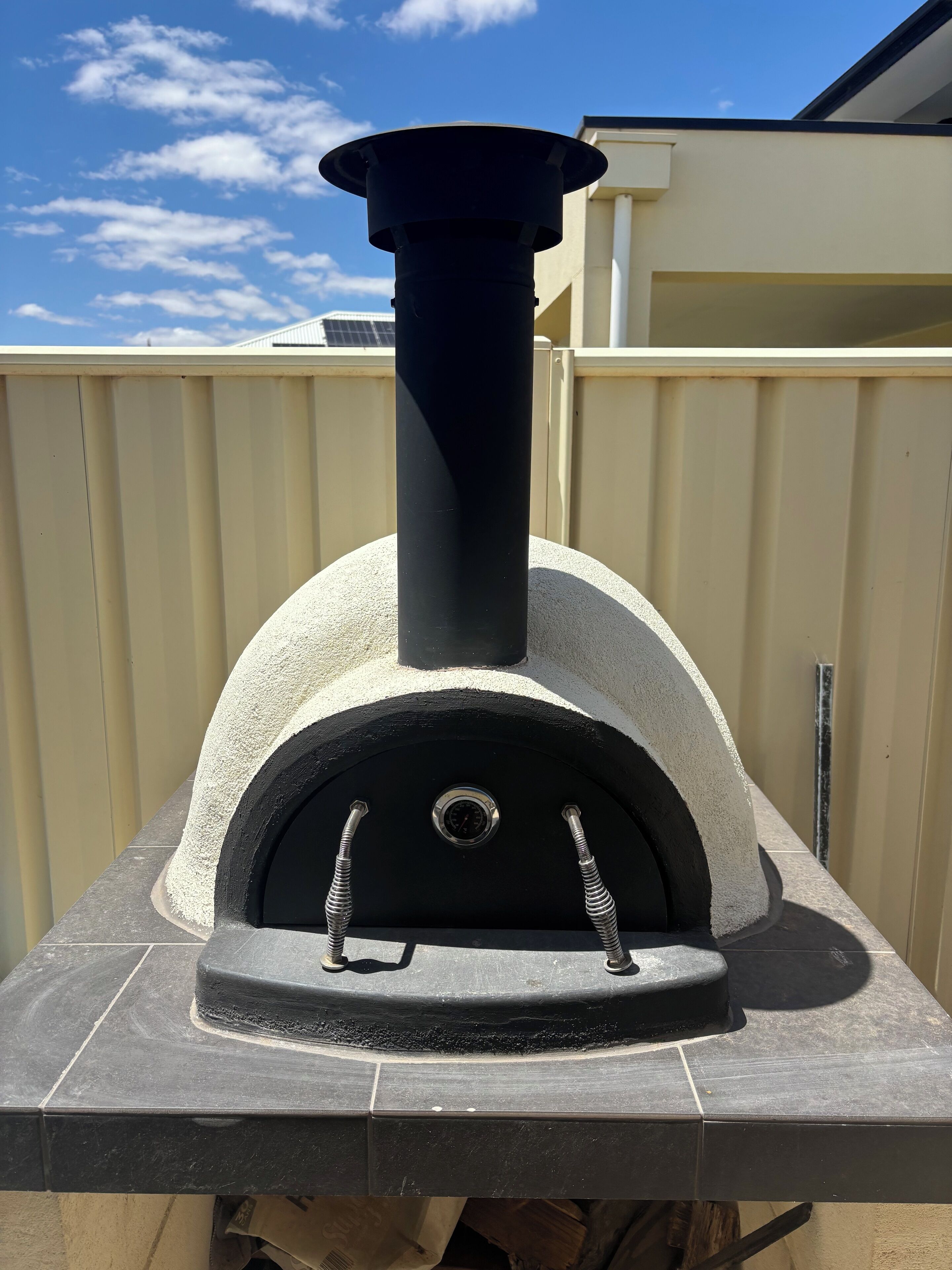Un four à pizza doté d’une cheminée noire et d’une façade en pierre, installé sur une plateforme carrelée.