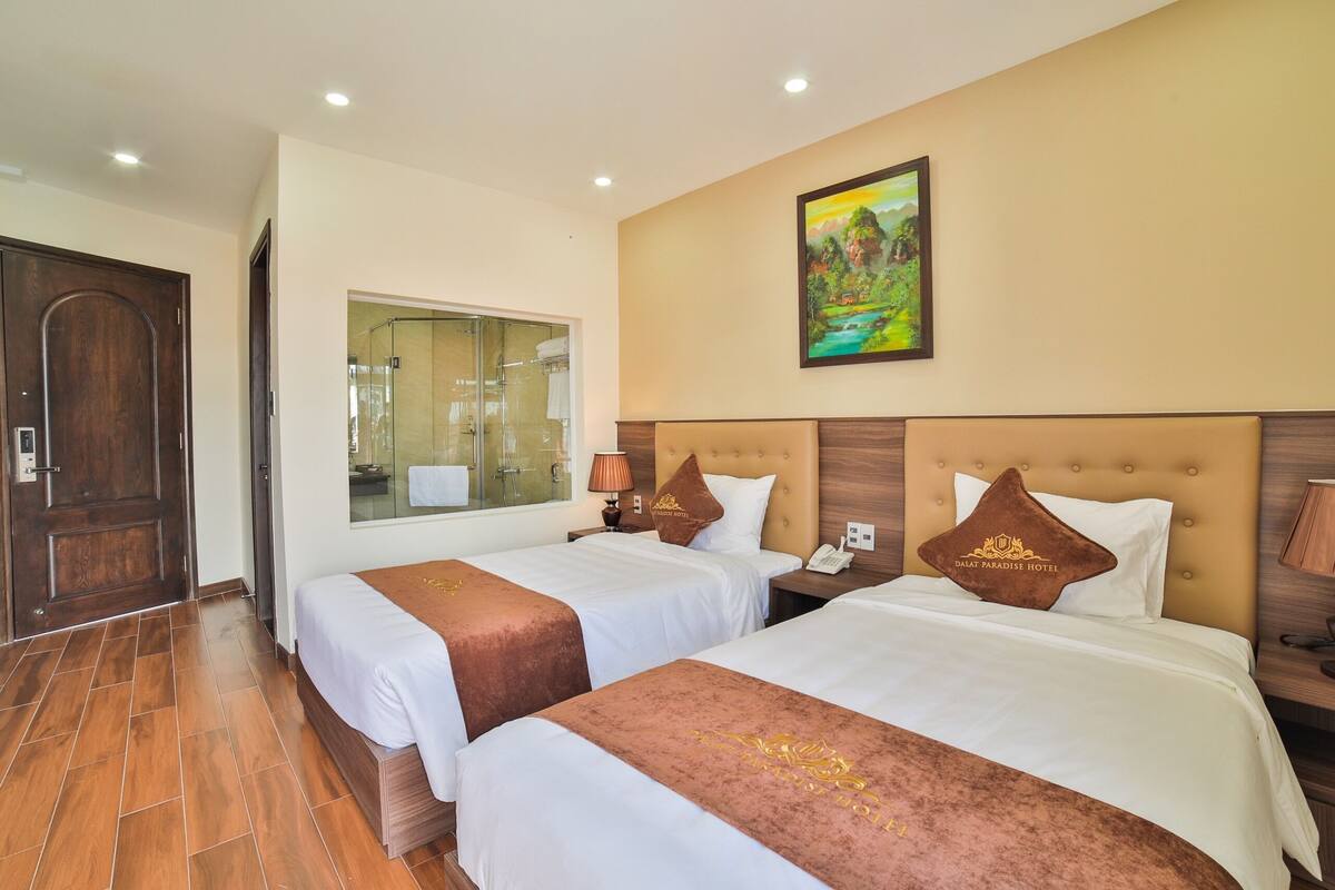 A hotel room with two beds, a wooden door, a glass shower area, and a painting on the wall.
