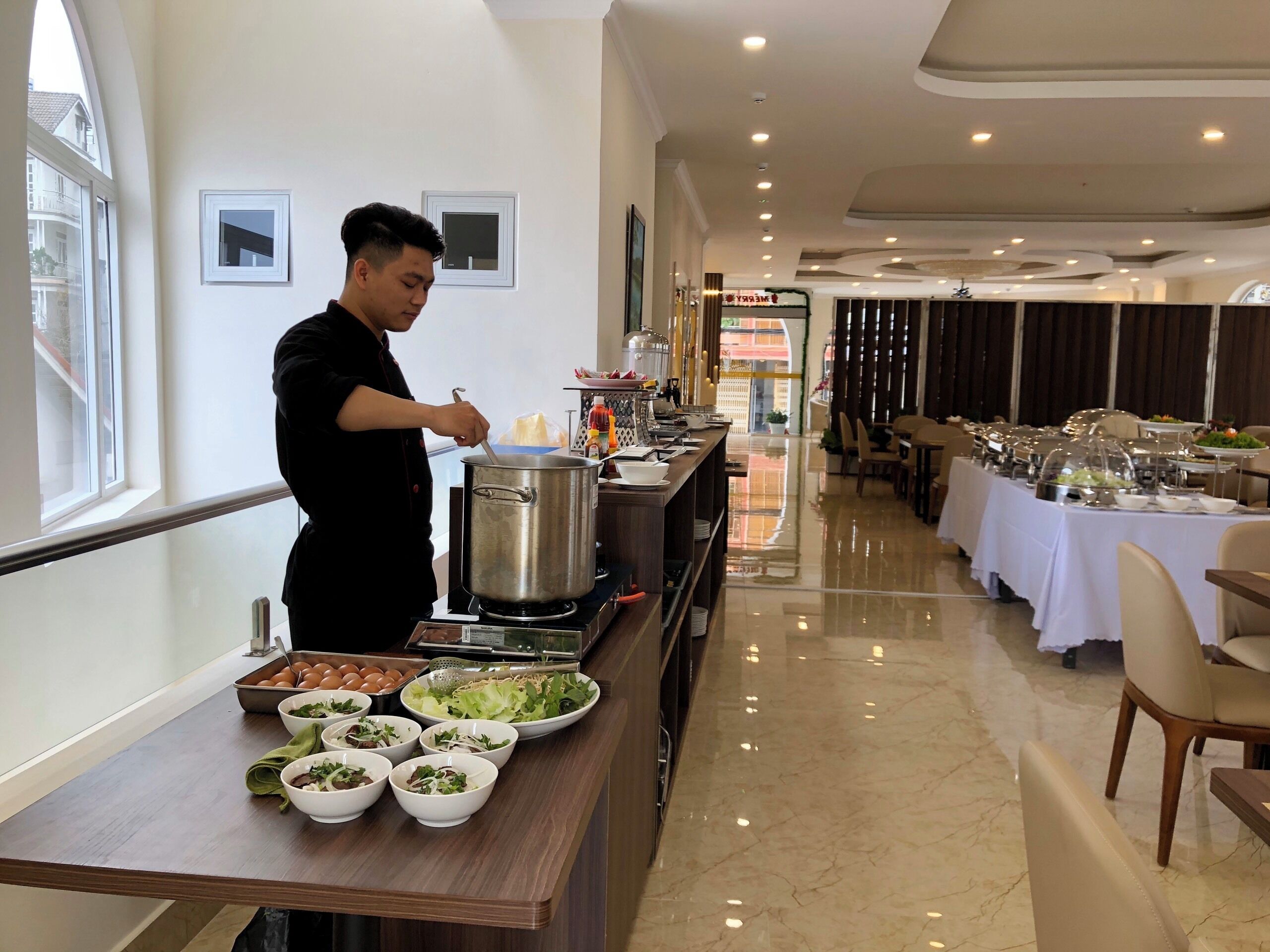 A hotel dining area with a chef preparing food, a buffet with various dishes, and a long table with chairs.