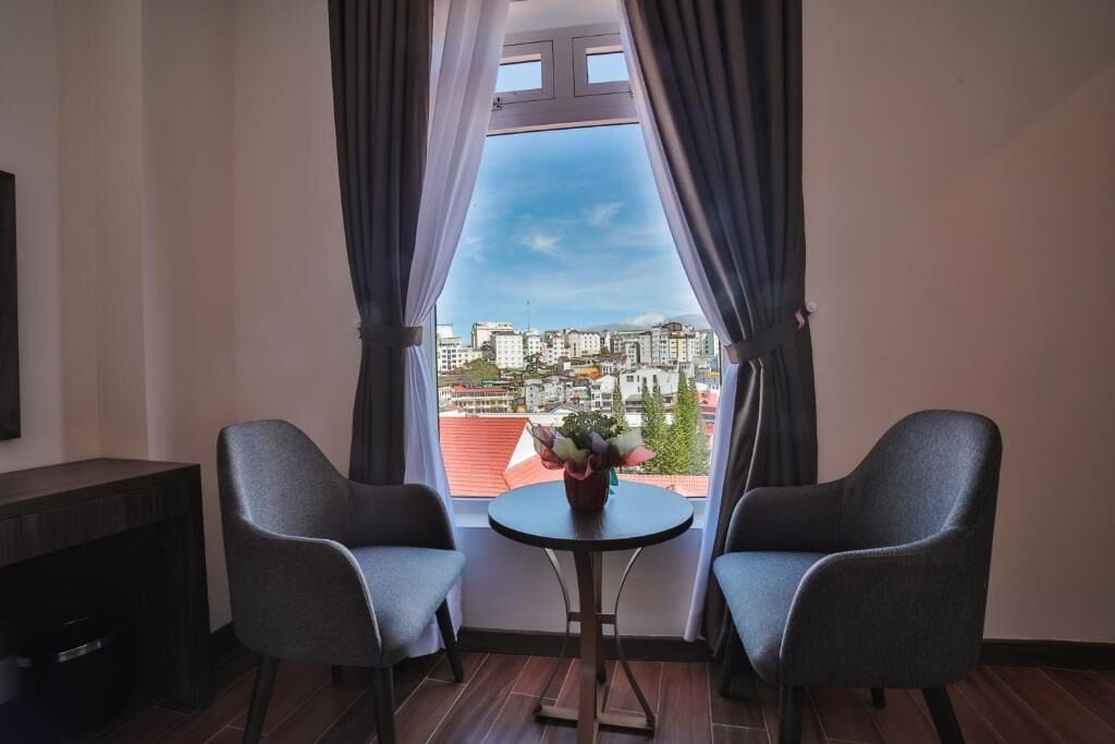 Una stanza con due poltrone grigie, un tavolino rotondo con un vaso di fiori e vista sulla città attraverso una finestra con tende.