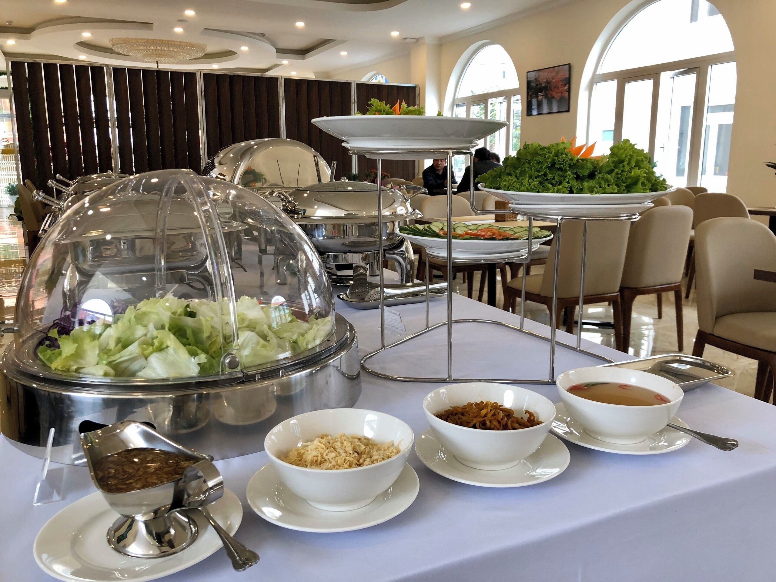 Montaje de bufé con estaciones de comida cubiertas, que incluyen ensalada, sopa y otros platos, en un área de comedor bien iluminada con mesas y sillas.