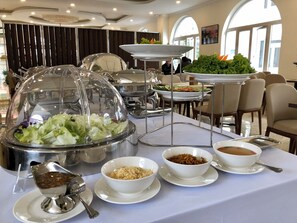 A buffet setup with covered food stations, including a salad, soup, and other dishes, in a well-lit dining area with tables and chairs. - Dalat Paradise Hotel (Xuan Huong - Da Lat)