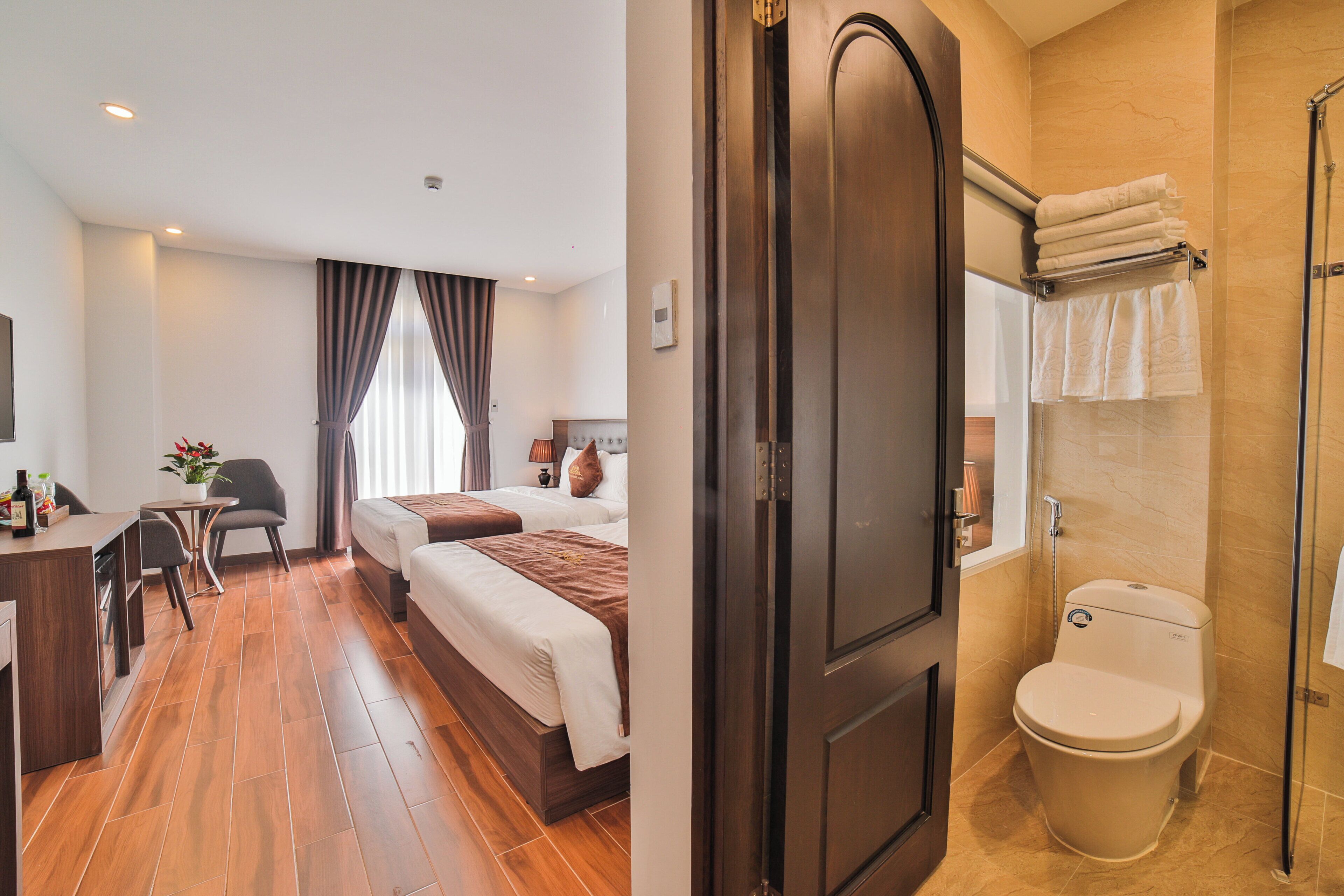 A hotel room with a wooden floor, a desk with a chair, two beds, a window with curtains, and a bathroom with a toilet and towel rack.