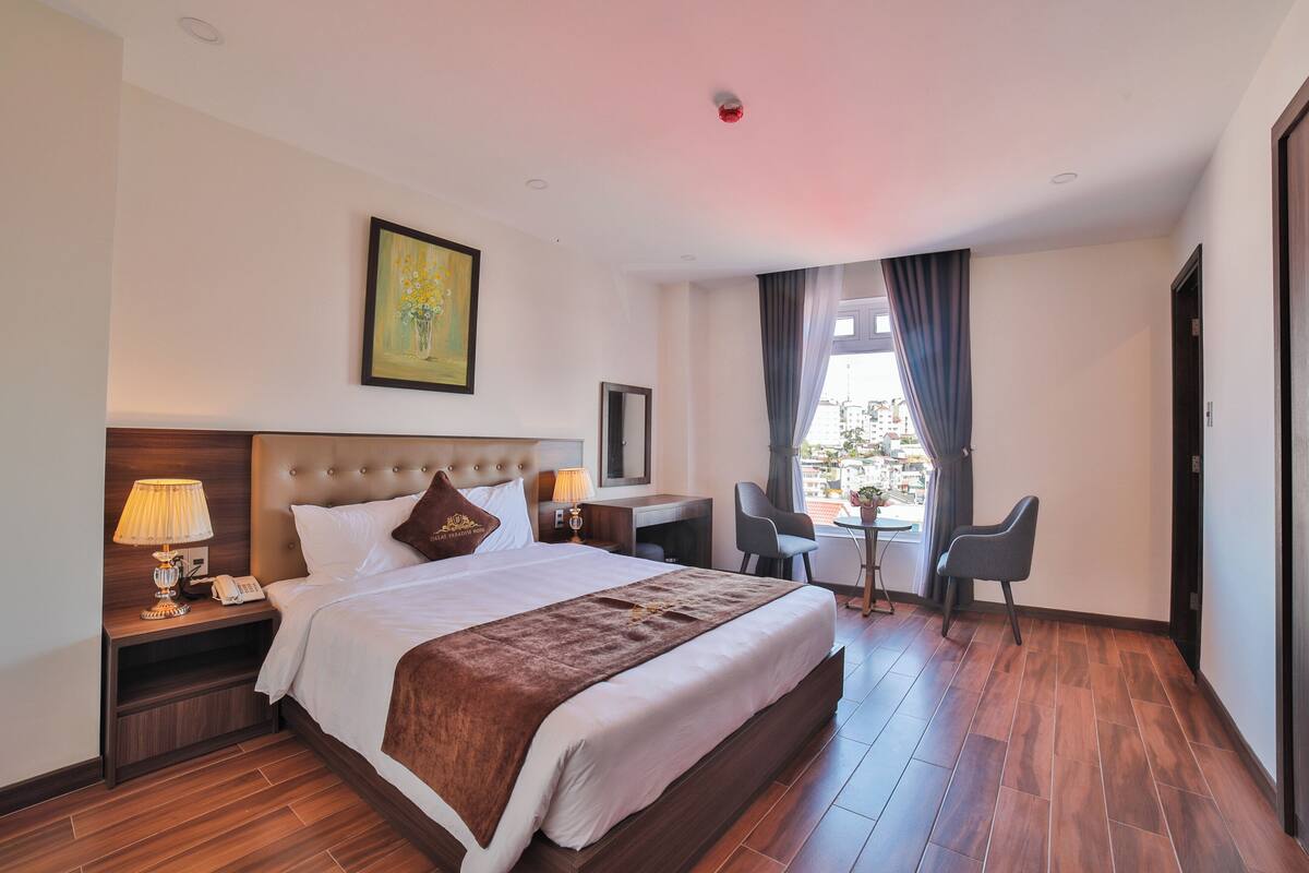 A hotel room with a large bed, a desk with a chair, a small table, and a window with curtains.