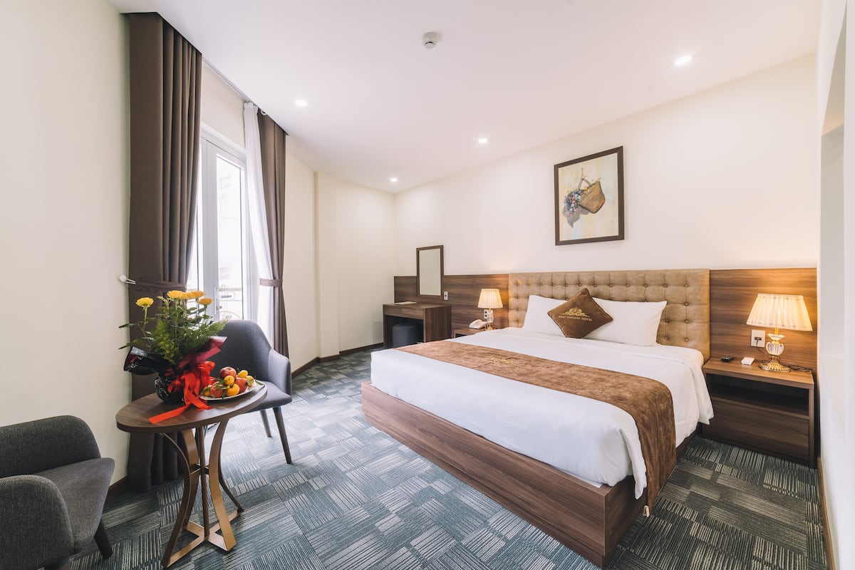 A hotel room with a large bed, a bedside table with lamps, a chair, a small table with a vase of flowers, and a painting on the wall.