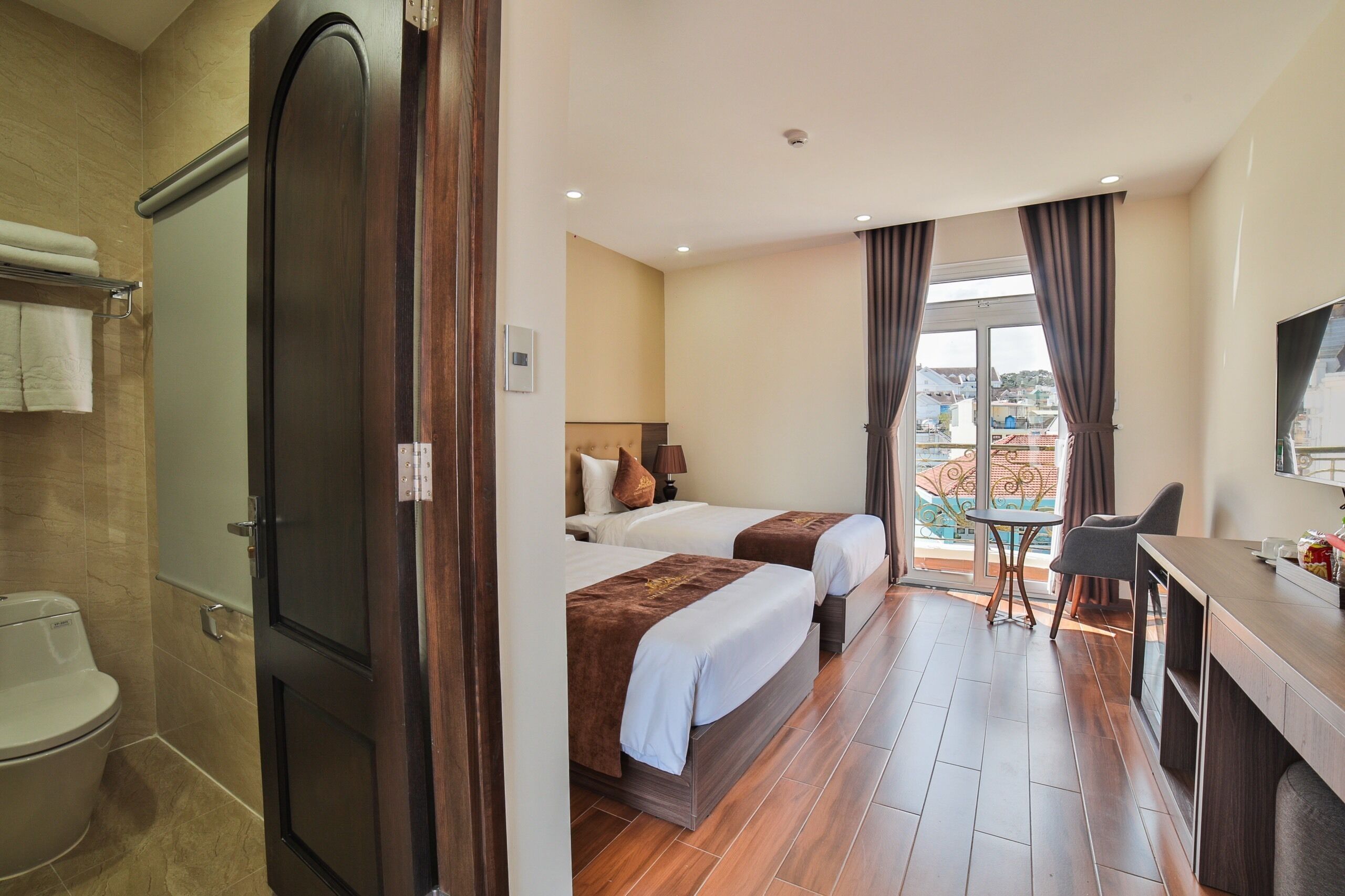Ein Hotelzimmer mit zwei Betten, einem Schreibtisch, einem Fernseher und einem Balkon mit Blick auf die Stadt.