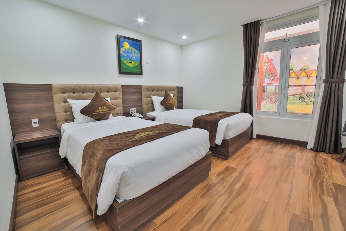 A hotel room with two beds, wooden headboards, a painting on the wall, and a window with curtains.