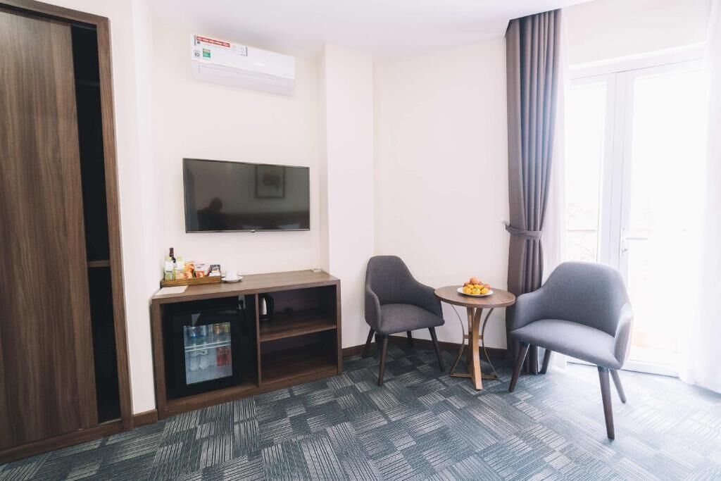 Habitación de hotel con televisión de pantalla plana en la pared, una mesita de madera con frutero, dos sillones grises, un mini refrigerador y piso alfombrado.