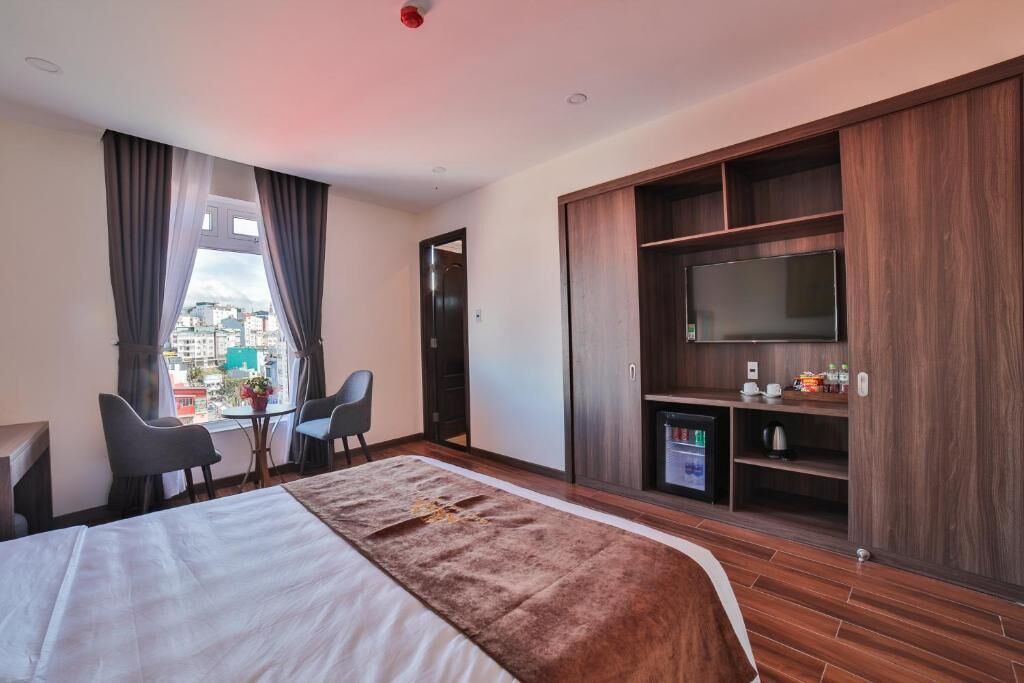 Habitación de hotel con cama, televisor, escritorio con sillas, una pequeña mesa con planta y vistas a la ciudad a través de la ventana.