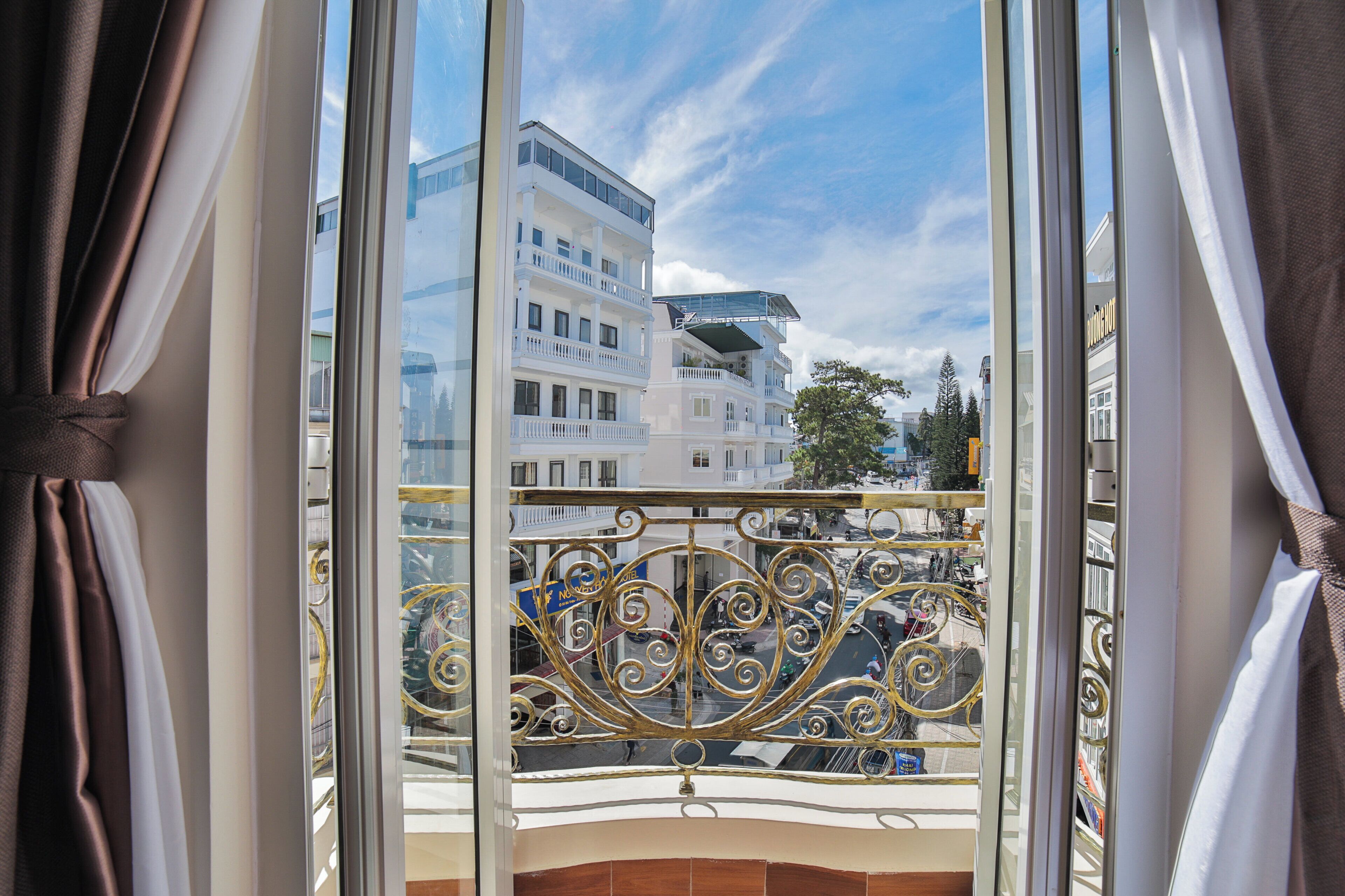 A balcony with ornate metal railings, overlooking a street with buildings and a clear sky.