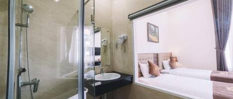 A hotel room with a glass shower enclosure, a sink, a hairdryer, and a bed with a headboard.