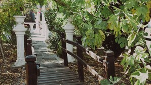 Uma passagem de madeira serpenteia por um jardim com colunas brancas e uma cerca de madeira.