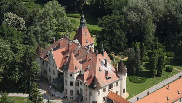 Vista aérea de um edifício histórico com uma torre central, rodeado por vegetação exuberante e um jardim bem cuidado.