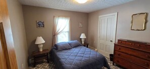 Une chambre à coucher avec un lit, deux tables de chevet avec des lampes, une commode, une porte et une fenêtre avec des rideaux.