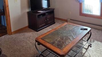 Una habitación con ventilador de techo, televisor sobre un mueble de madera oscura, alfombra estampada y una ventana con cortinas azules.