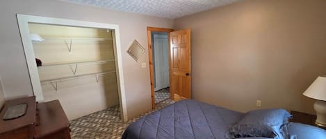 A bedroom with a bed, a dresser, a door leading to another room, and a wall-mounted shelf.