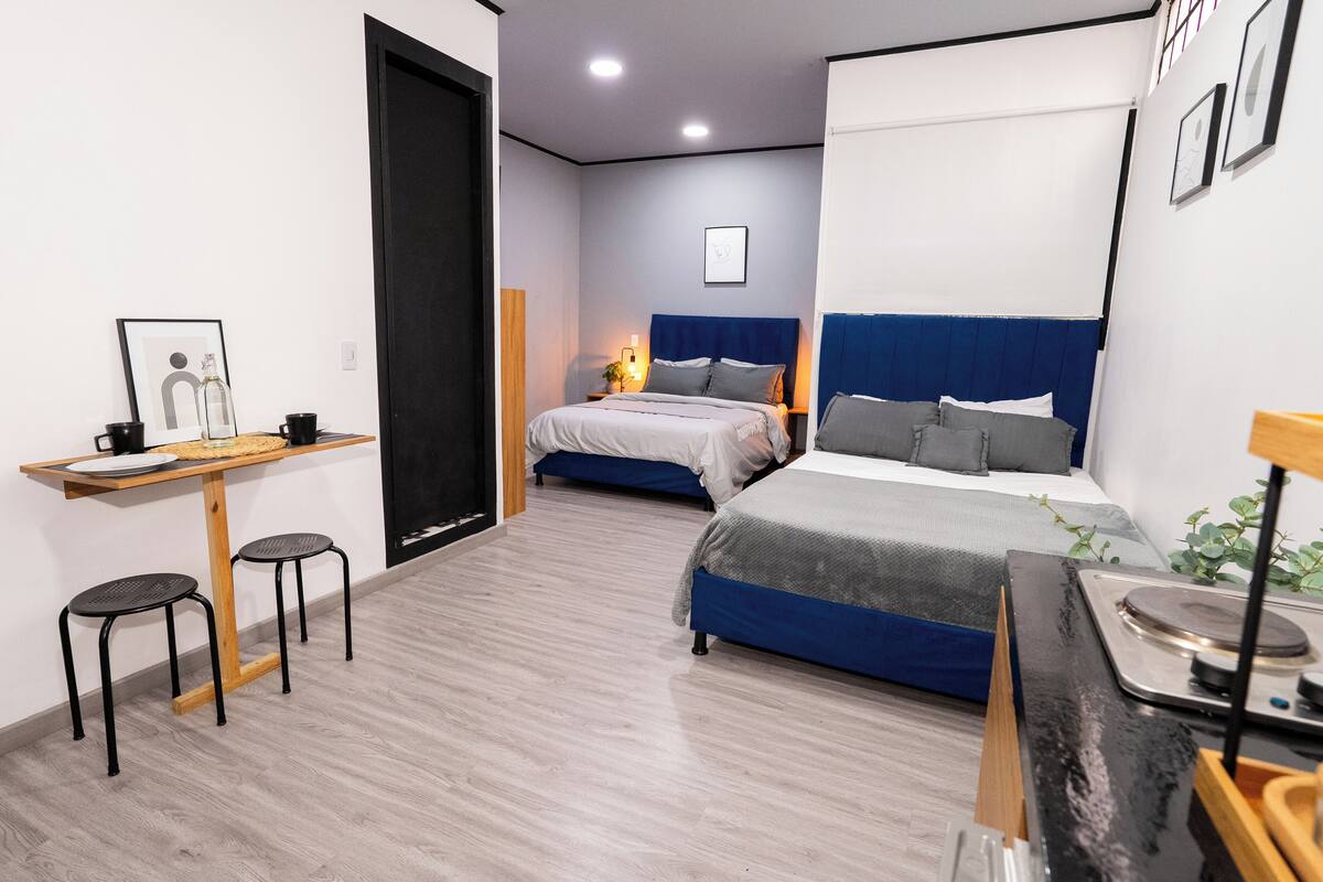 Une chambre d’hôtel moderne avec un bureau en bois, deux tabourets noirs, un lit avec une tête de lit bleue et une petite table avec une lampe.
