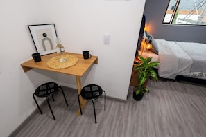 Une petite pièce avec un bureau en bois, deux chaises noires, un tableau encadré, une tasse noire, un set de table en fibres tressées, une plante en pot et un lit recouvert d’une couverture blanche.