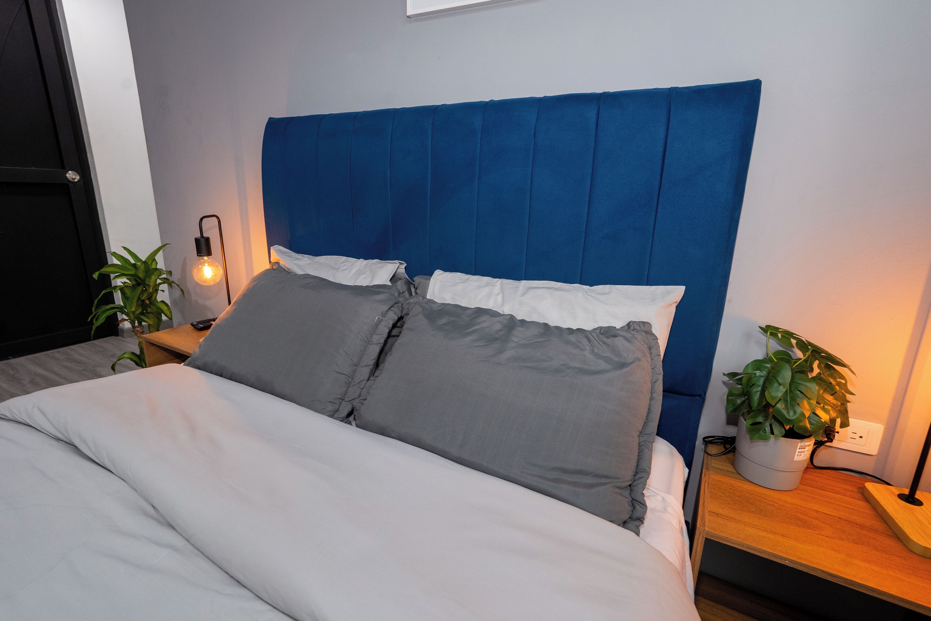 Una cama bien hecha con un cabecero azul, una lámpara de noche y una planta en maceta sobre una mesita de madera.