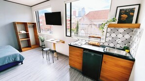 Une chambre d’hôtel compacte comprenant un lit, une petite kitchenette, un coin repas et une grande fenêtre donnant sur la ville.