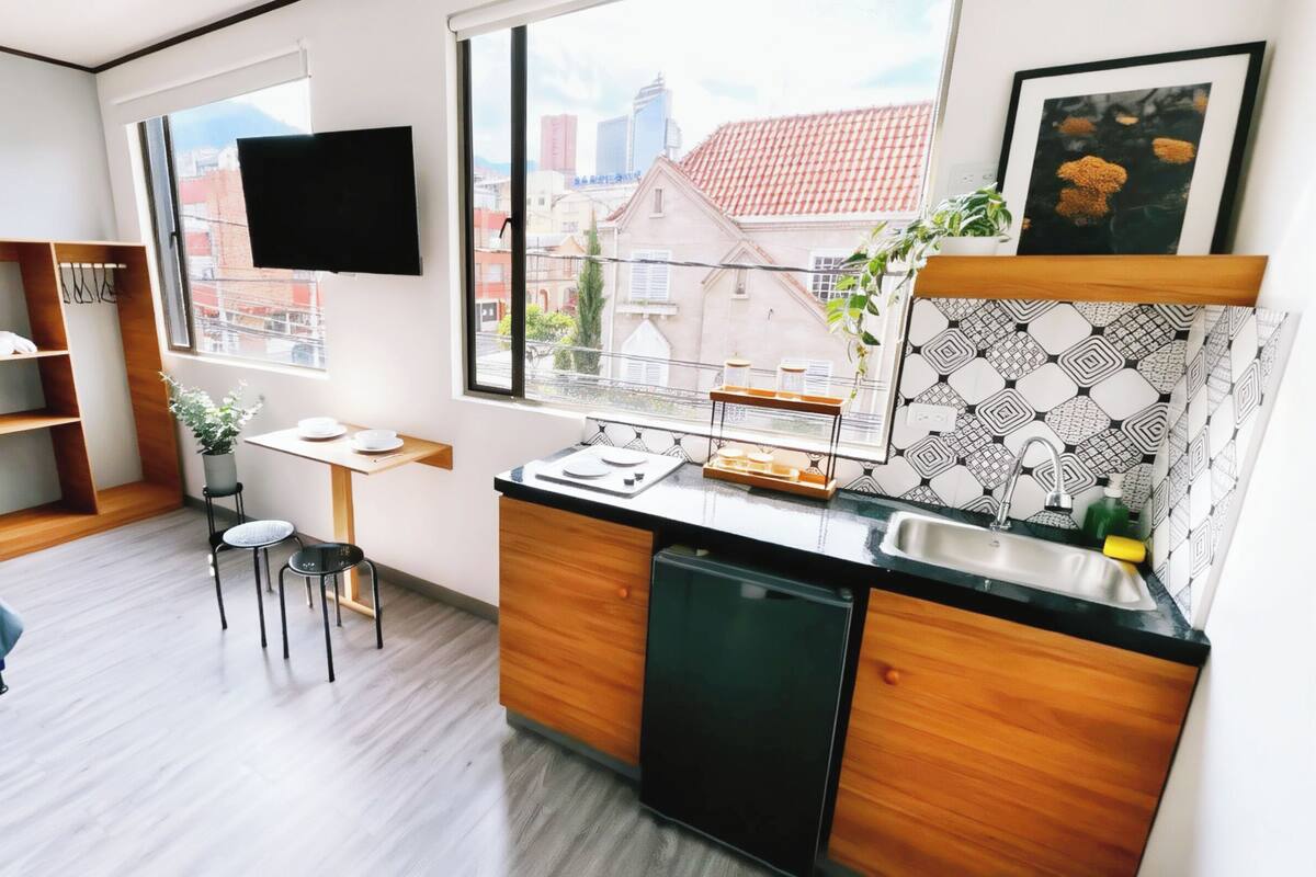 Une chambre d’hôtel compacte comprenant un lit, une petite kitchenette, un coin repas et une grande fenêtre donnant sur la ville.