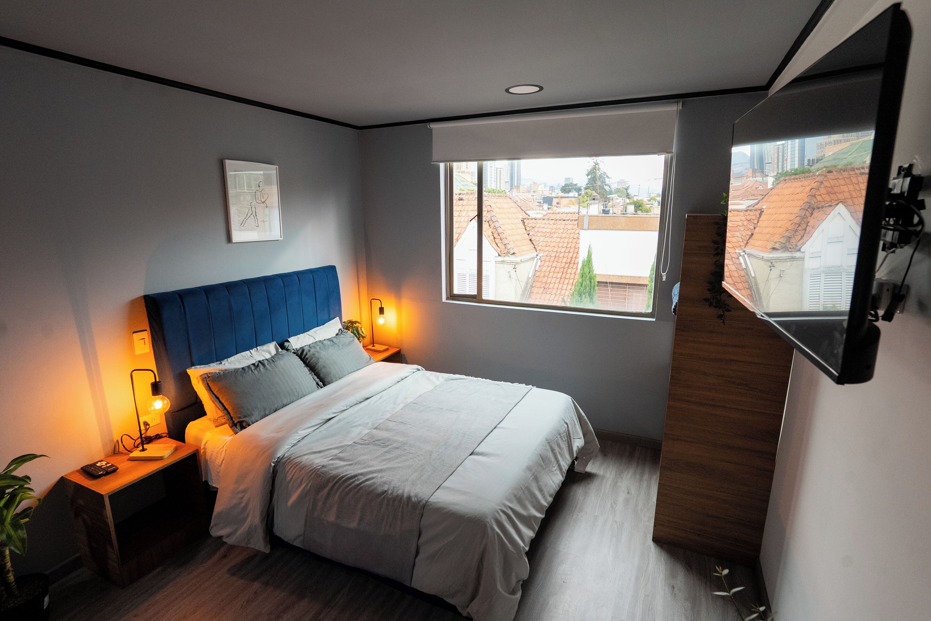 A bedroom with a bed, bedside lamps, a window with a view of buildings, and a mounted TV.