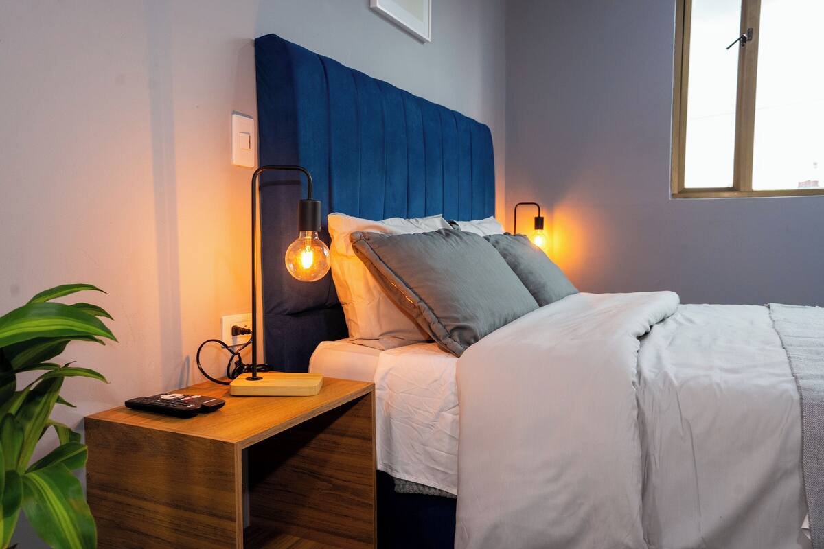 Un lit bien fait, avec une tête de lit bleue, une table de chevet avec une lampe et une plante.