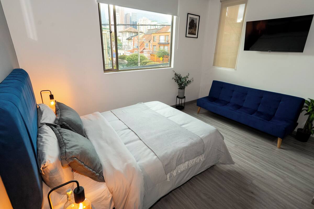 Une chambre moderne avec un lit, un canapé bleu, une télévision et une fenêtre donnant sur la ville.
