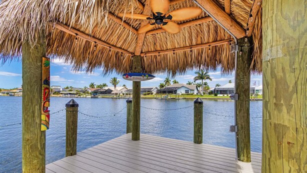 A wooden dock with a thatched roof and a ceiling fan, overlooking a body of water with houses in the background. - Riverfront Escape with Pool, Tiki Hut & Gulf Access Fun - Villa Kai Malie - Roelens (Cape Coral)