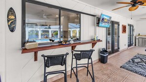 A bar area with a large window, two black wicker chairs, a television, and a ceiling fan. - Riverfront Escape with Pool, Tiki Hut & Gulf Access Fun - Villa Kai Malie - Roelens (Cape Coral)