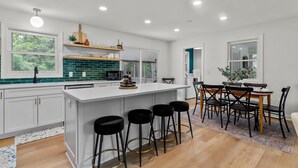 A modern kitchen with white cabinetry, a green backsplash, and a central island with stools. - The Charming Pines Hideaway - 3B/2B - Golfer's Paradise (Southern Pines)