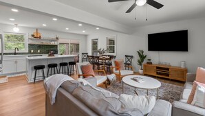 A modern kitchen with white cabinets, a dining area with a table and chairs, a living room with a sofa and a TV mounted on the wall. - The Charming Pines Hideaway - 3B/2B - Golfer's Paradise (Southern Pines)