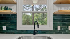 A kitchen with a window, green tiles, a stainless steel sink, and wooden shelves. - The Charming Pines Hideaway - 3B/2B - Golfer's Paradise (Southern Pines)