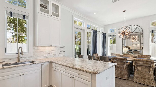 A modern kitchen with white cabinetry, a granite countertop, and a dining area with wicker furniture. - Edge of Rosemary Beach + 1 Minute Walk to Beach | Pool, Putting Green, Carriage House (Rosemary Beach)