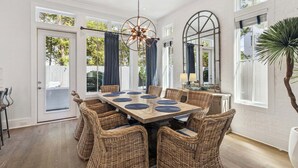 A dining room with a large table, wicker chairs, a chandelier, and large windows. - Edge of Rosemary Beach + 1 Minute Walk to Beach | Pool, Putting Green, Carriage House (Rosemary Beach)