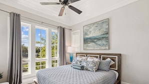 A bedroom with a large bed, a ceiling fan, a window with curtains, a painting on the wall, and a nightstand with a lamp. - Edge of Rosemary Beach + 1 Minute Walk to Beach | Pool, Putting Green, Carriage House (Rosemary Beach)