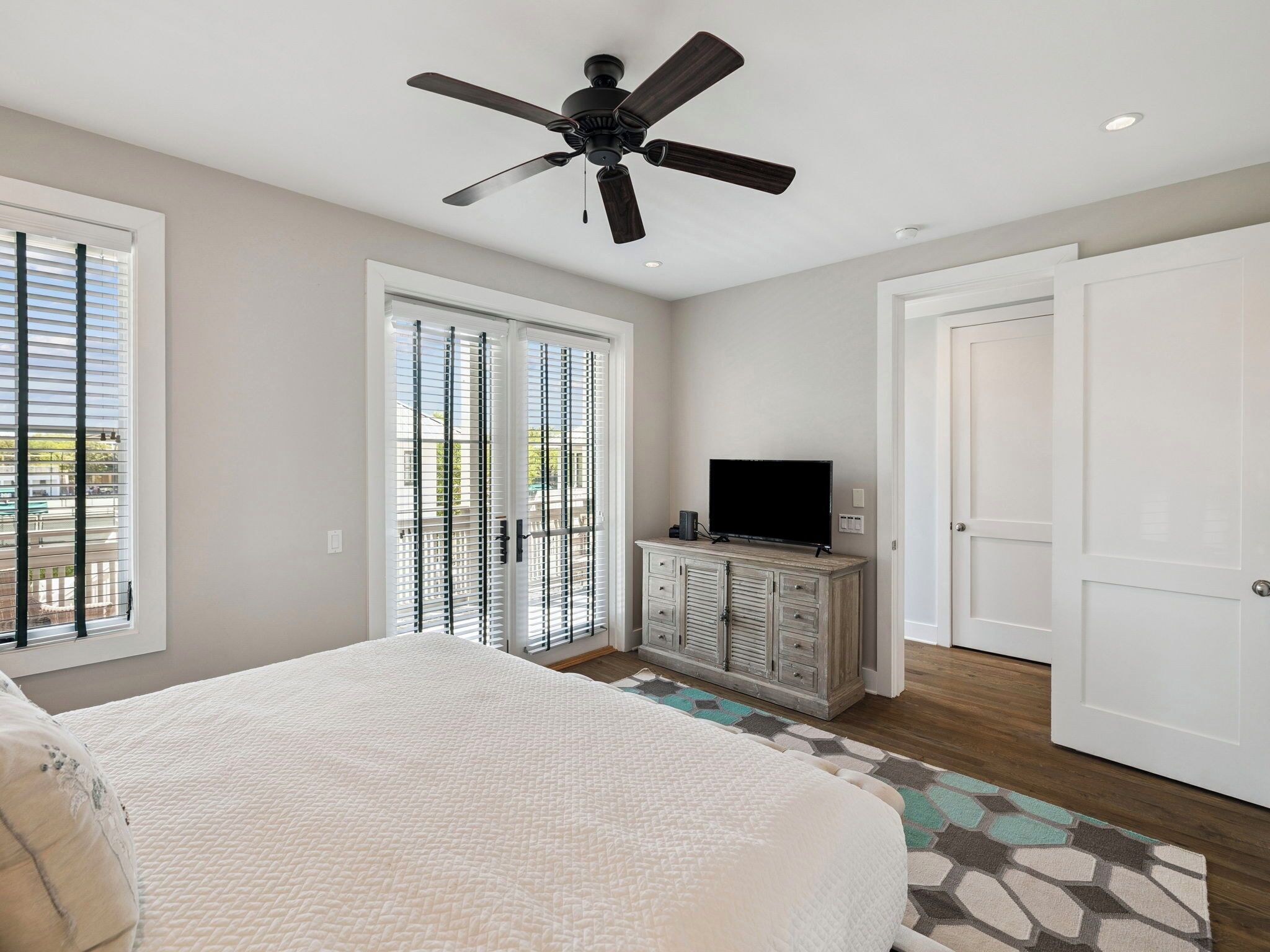Une chambre à coucher avec un lit, un ventilateur de plafond, un téléviseur posé sur une commode et une porte-fenêtre coulissante.