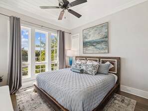 Un dormitorio con una cama grande, un ventilador de techo, una ventana con cortinas, un cuadro en la pared y una mesita de noche con una lámpara.